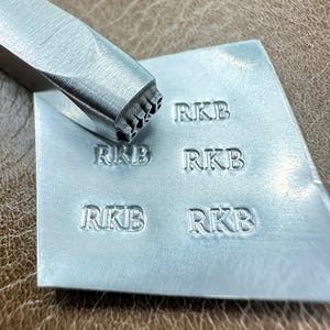 May include: A metal stamp tool with the letters "RKB" stamped repeatedly onto a silver metal surface. The tool is silver and angled, with the letters clearly visible. The background is a brown leather-like material.
