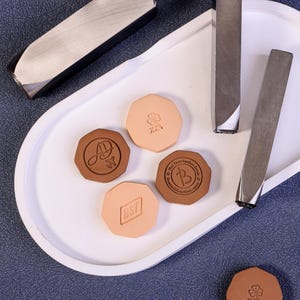 May include: A collection of leather craft tools and stamped leather pieces. The tools are silver-colored, and the leather pieces are in shades of tan and brown. Some pieces have stamped designs and text.