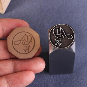 Könnte beinhalten: Ein brauner achteckiger Wachssiegel und ein Metallstempel mit rundem Design. Der Siegel zeigt die Buchstaben "AD" und ein kleines Flugzeug. Der Stempel hat das gleiche Design mit den Buchstaben "AD" und einem kleinen Flugzeug.