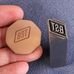 Könnte beinhalten: Zwei Metallstempel, einer achteckig und einer rechteckig, mit den Buchstaben "BST" und "T8B". Der achteckige Stempel ist beige, und der rechteckige Stempel ist dunkelgrau mit silbernem Rand. Die Stempel werden wahrscheinlich zum Prägen verwendet.