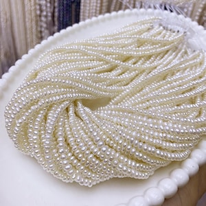 Puede incluir: Colección de pequeñas perlas redondas de color blanco cremoso, ensartadas en múltiples hilos. Las perlas están dispuestas en un patrón arremolinado sobre una bandeja blanca con borde festoneado. Las perlas tienen un brillo sutil.