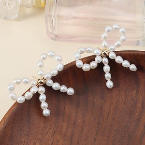 Può includere: Un paio di orecchini a forma di fiocco. Ogni orecchino è realizzato con perle bianche di forma irregolare, disposte a formare un fiocco. Un piccolo elemento metallico dorato fissa le perle al centro. Gli orecchini sono esposti su una superficie di legno.