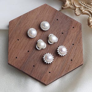 May include: Six pearl earrings in different designs are presented on a wooden hexagonal stand. The earrings feature white pearls with silver-coloured settings, some with a halo of small, clear stones. The wood has a rich, dark brown colour.
