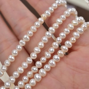 Pode incluir: Close-up de vários fios de colares de pérolas brancas. As pérolas são pequenas e têm uma forma ligeiramente irregular. Os colares são exibidos em um fundo neutro.