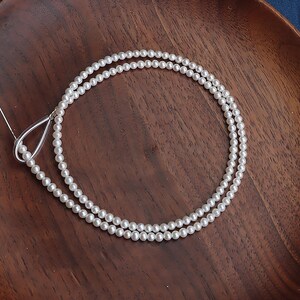 May include: A delicate, double-strand pearl necklace with a silver teardrop-shaped clasp. The small, round pearls are white and evenly spaced. The necklace is displayed on a dark wooden surface.