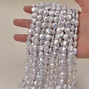 Puede incluir: Cadenas de perlas blancas en forma de moneda. Las perlas tienen un brillo iridiscente sutil, que refleja toques de rosa y azul. Las cuentas están ensartadas, creando una apariencia fluida y texturizada. Ideal para la creación de joyas.