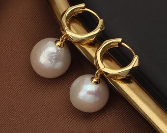 Puede incluir: Un par de pendientes de aro dorados con grandes perlas blancas redondas. Los pendientes tienen un diseño geométrico en los aros y un pequeño conector dorado que sujeta la perla. El fondo es una mezcla de marrón y negro.