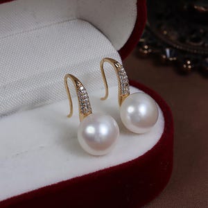 May include: A pair of gold drop earrings, each with a large, round, white pearl. The top section of each earring has a curved design with small, sparkling embellishments. The earrings are presented in a red velvet jewellery box.
