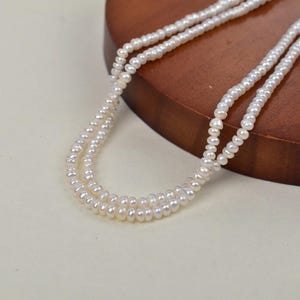 May include: A double-strand pearl necklace. The necklace is made of small, round, off-white pearls. The strands are draped over a wooden surface. The background is a soft, neutral colour.