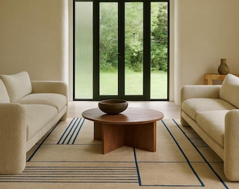 Könnte beinhalten: Ein modernes Wohnzimmer mit zwei beigefarbenen Sofas, einem runden Couchtisch aus Holz und einem gemusterten Teppich mit blauen Linien. Ein großes Fenster bietet einen Blick auf einen grünen Außenbereich. Der Text "therugfusion" ist auf dem Teppich zu sehen.