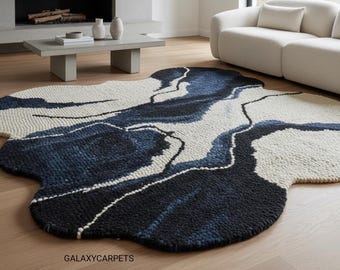 Op de afbeelding: Een groot, abstract tapijt met een unieke vorm en een ontwerp dat lijkt op een stromende rivier. Het tapijt heeft tinten marineblauw, zwart en wit, wat een moderne en artistieke uitstraling creëert. Het tapijt ligt op een lichtgekleurde houten vloer.