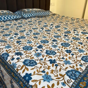 Op de afbeelding: Een witte sprei en twee kussenslopen met een blauw bloemenpatroon en bruine wijnranken. De sprei heeft een blauwe rand met een herhalend geometrisch ontwerp. De kussens hebben een vergelijkbaar patroon. Het beddengoed ligt op een bed met een bruin hoofdbord.