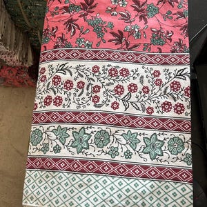 Puede incluir: Tela doblada con un estampado floral. La sección superior es rosa liso con flores verdes y blancas. Debajo hay bandas florales blancas y burdeos, y un patrón de diamantes verdes y blancos en la parte inferior. La tela está doblada.