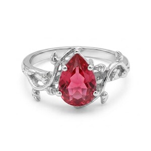 Könnte beinhalten: Ein silberner Ring mit einem birnenförmigen, facettierten roten Edelstein in der Mitte. Die Ringschiene ist mit einem strukturierten, rankenartigen Motiv gestaltet, das sich um die Seiten des zentralen Steins windet und kleine Blattdetails enthält.