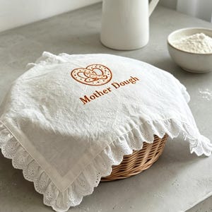 May include: A white linen bread basket cover with a decorative scalloped edge and embroidered pretzel design. The words "Mother Dough" are embroidered in brown. The cover is placed over a wicker basket.