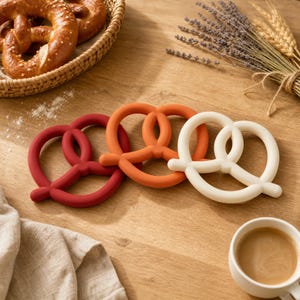 May include: Three pretzel-shaped silicone items in red, orange, and cream colors arranged in a row. These smooth, circular objects feature the classic twisted knot design of a pretzel, providing a solid, matte appearance for each individual piece.