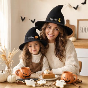 May include: Two black pointed witch hats featuring custom text and icons. One hat displays the name Emily with an orange jack-o'-lantern, while the other hat features the name Olivia with a yellow crescent moon and a small star icon.