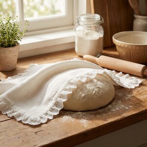 May include: A square off-white cloth with a delicate lace-trimmed ruffled edge draped over a round mound of raw bread dough. The fabric features a smooth texture and intricate eyelet detailing along the scalloped border of the trim.