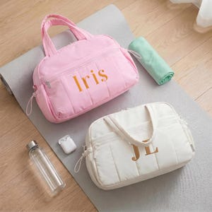Könnte beinhalten: Eine hellrosa gesteppte Reisetasche mit dem in Gold gestickten Namen Iris und eine weiße gesteppte Reisetasche mit den in Gold gestickten Initialen JL. Beide Taschen verfügen über Tragegriffe, Reißverschlusstaschen vorne und passende Zipper.