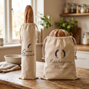 Puede incluir: Dos bolsas de pan de lona beige con cierres de cordón. Una bolsa contiene una baguette, la otra un pan redondo. La bolsa de la izquierda tiene el texto "Emma's Baguette" y un gráfico de trigo. La bolsa de la derecha tiene el texto "Boulangerie Maison" y un gráfico de trigo.