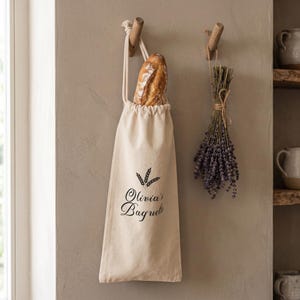 May include: A beige fabric drawstring bag hanging from a wooden hook, containing a crusty baguette. The bag features a black graphic of wheat stalks and the text "Olivia's Baguette" printed on the front. A bundle of dried lavender hangs nearby.