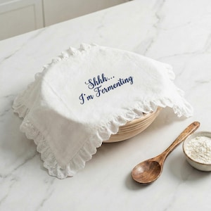 Puede incluir: Paño de lino blanco para fermentación de pan con borde con volantes, que cubre una cesta redonda. El texto "Shhh... I'm Fermenting" está impreso en azul marino. Una cuchara de madera y un cuenco pequeño con harina están junto a la cesta.