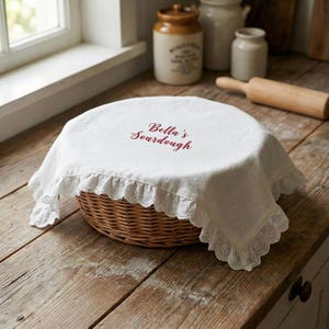May include: A round, woven wicker basket covered with a white cloth. The cloth has a scalloped lace trim and the words "Bella's Sourdough" in burgundy script. The basket sits on a rustic wooden surface.