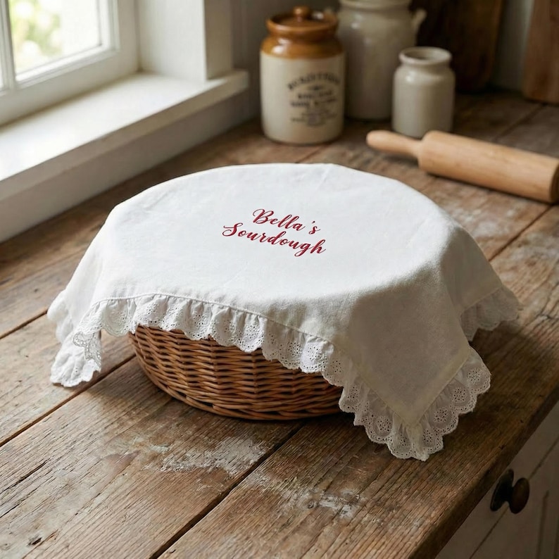 May include: A round, woven wicker basket covered with a white cloth. The cloth has a scalloped lace trim and the words "Bella's Sourdough" in burgundy script. The basket sits on a rustic wooden surface.