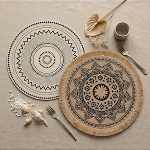 May include: Two round placemats with black geometric patterns. One is white with a circular mandala-style design. The other is tan with a fringed edge and a bold, dark, intricate circular pattern. Two silver forks, a small cup, and seashells are present.