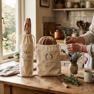 May include: Two beige drawstring bread bags with black text and illustrations. One bag holds a baguette, the other a round loaf. The text on the bags reads "Le Pain Frais" and "Boulangerie Maison". The bags sit on a wooden table.