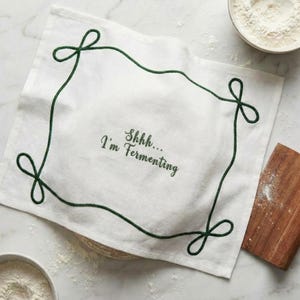 May include: White linen kitchen towel with a green embroidered border and the text "Shhh... I'm Fermenting". The towel has a rectangular shape with a decorative border and is likely used for baking or cooking.