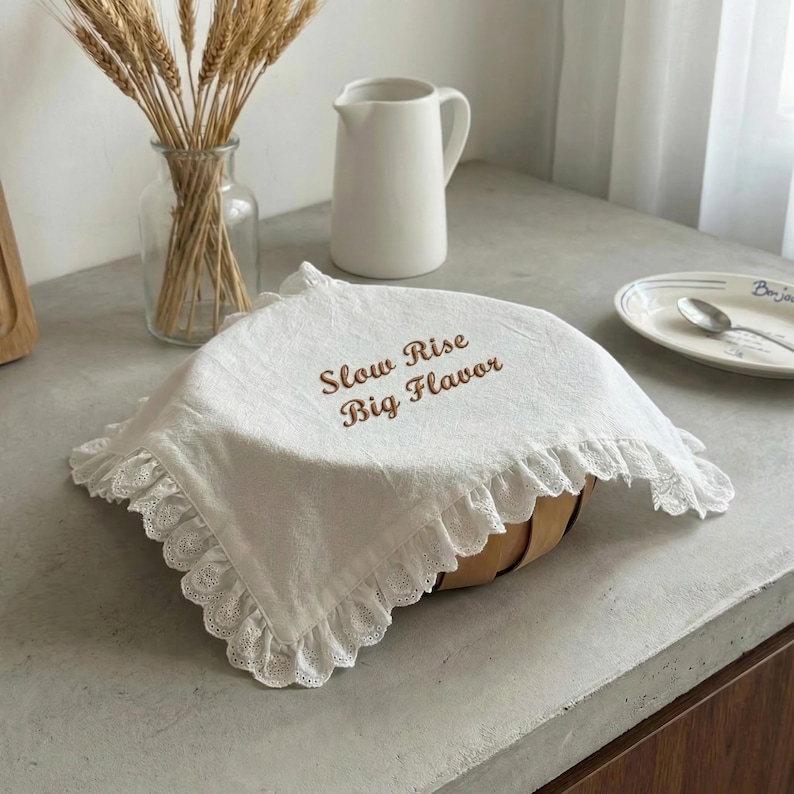 May include: A white, ruffled bread cover with the text "Slow Rise Big Flavor" in brown script. The cover is draped over a basket. A clear glass vase with wheat stalks, a white pitcher, and a plate with a spoon are in the background.