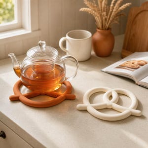 May include: A clear glass teapot filled with amber tea sits on a burnt orange pretzel-shaped silicone trivet. Beside it lies an identical cream-coloured pretzel-shaped silicone trivet. The items are arranged on a light-coloured countertop.
