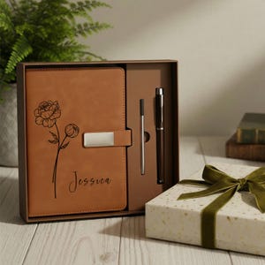 May include: A brown leather journal with a floral design and the name "Jessica" is displayed in a gift box with a pen and a ballpoint pen. A gift box with a green ribbon is also visible.