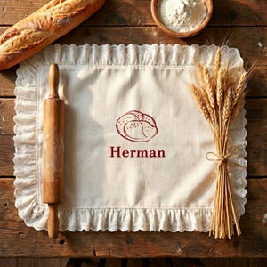 May include: A rustic scene featuring a cream-colored placemat with a ruffled lace edge. A wooden rolling pin and a loaf of bread are on the left. The placemat has a bread illustration and the name "Herman" printed in red. A bundle of wheat and a bowl of flour are on the right.