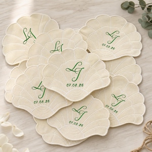 May include: A collection of cream-coloured fabric coasters shaped like seashells. Each coaster features green embroidered monogram initials LT and the date 07.08.26. The fabric has a quilted texture with stitched lines radiating from the centre.