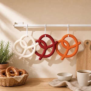 May include: Three pretzel-shaped trivets in white, dark red, and orange hang from a white horizontal rod. Each trivet features a looped design and is suspended by a white hook, providing a functional kitchen accessory for heat protection.