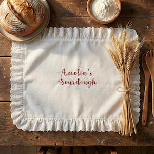 May include: A white placemat with a ruffled lace edge, embroidered with "Amelia's Sourdough" in red script. A loaf of bread sits on a plate, with a bundle of wheat and wooden utensils nearby. A bowl of flour is also present.