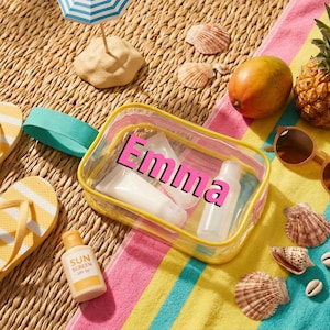 May include: A clear, personalized travel cosmetic bag with the name "Emma" in pink, containing white bottles and tubes. The bag is on a striped towel with a mango, pineapple, sunglasses, seashells, and sunscreen.