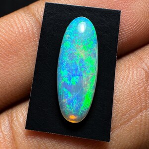 May include: An oval-shaped cabochon opal gemstone featuring a translucent body with vibrant flashes of neon green and electric blue play-of-color, set against a small rectangular black backing card for contrast and display.