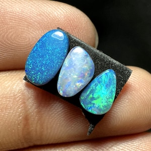 Könnte beinhalten: Drei polierte Opal-Edelsteine in Blau- und Grüntönen. Die Opale sind oval und tropfenförmig, mit schillernden Mustern. Die Edelsteine werden auf einer schwarzen Oberfläche präsentiert, die zwischen den Fingern gehalten wird.