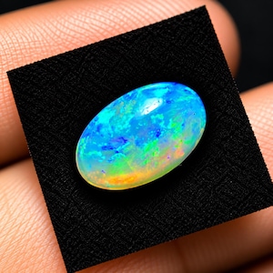 May include: An oval-shaped loose gemstone featuring a vibrant play-of-color with dominant shades of electric blue, bright green, and subtle hints of orange and yellow, set against a solid black square background for high contrast.