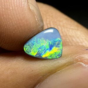 May include: A polished, triangular opal gemstone. The opal has a light blue base color with vibrant internal flashes of green, yellow, and blue. The stone is held between fingers, showcasing its iridescent play-of-color.