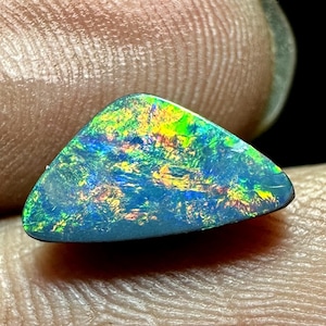 May include: A triangular-shaped black opal gemstone featuring a dark base color with vibrant flashes of green, blue, and yellow play-of-color across its polished surface. The stone exhibits a distinct iridescent pattern throughout its structure.