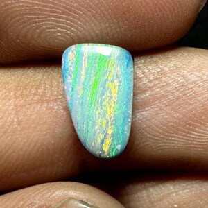 May include: A polished opal gemstone with a vibrant display of colors. The stone features a mix of blue, green, and yellow hues, with a play of light creating a shimmering effect. The opal is held between fingers, showcasing its unique shape and natural beauty.