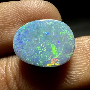 May include: A polished, oval-shaped opal gemstone. The stone displays a milky, light blue base color with vibrant flashes of green, yellow, and blue iridescence. The opal is held between fingers, showcasing its natural beauty.