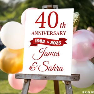 May include: A white sign on a wooden easel, celebrating a 40th anniversary. The sign features red text that reads "40th Anniversary 1985-2025 James & Sahra." Balloons in white, gold, and pink are in the background.
