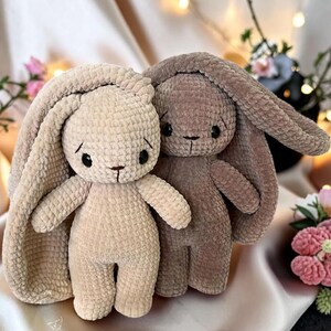 Könnte beinhalten: Zwei Plüschtiere in Form von Hasen mit langen Ohren. Ein Hase ist hellbeige und der andere dunkelbraun. Beide haben eine weiche, strukturierte Oberfläche aus gehäkeltem Garn, runde schwarze Augen sowie eine kleine gestickte Nase und Mund.