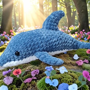 Könnte beinhalten: Ein Plüschdelfin aus strukturiertem blauem Garn mit weißem Bauch und einem glatten schwarzen Auge. Das Stofftier verfügt über eine markante Rückenflosse, Seitenflossen und einen Schwanz und zeigt ein weiches, gehäkeltes Erscheinungsbild am ganzen Körper.