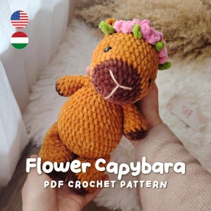 Puede incluir: Un juguete de peluche de capibara tejido a ganchillo en color marrón con hocico marrón oscuro, ojos negros y una corona de flores rosas con hojas verdes en la cabeza. El texto Flower Capybara PDF CROCHET PATTERN aparece abajo.